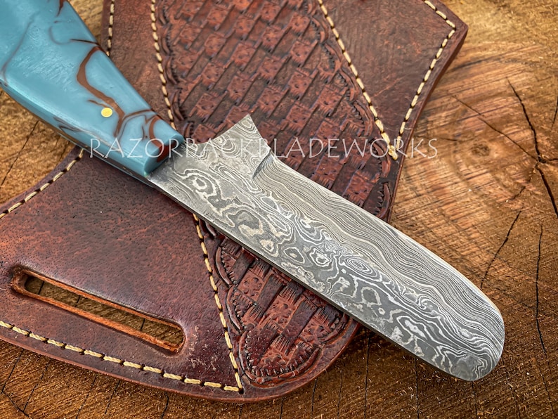 May include: A straight razor with a blue and brown marbled handle and a damascus steel blade. The razor is resting on a brown leather sheath with decorative stitching.