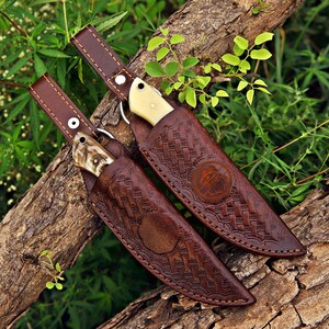 May include: Two fixed-blade knives with cream-colored handles, each housed in a dark brown leather sheath featuring an intricate basket-weave pattern, a circular embossed logo, and a long strap with a silver-toned metal snap button closure.
