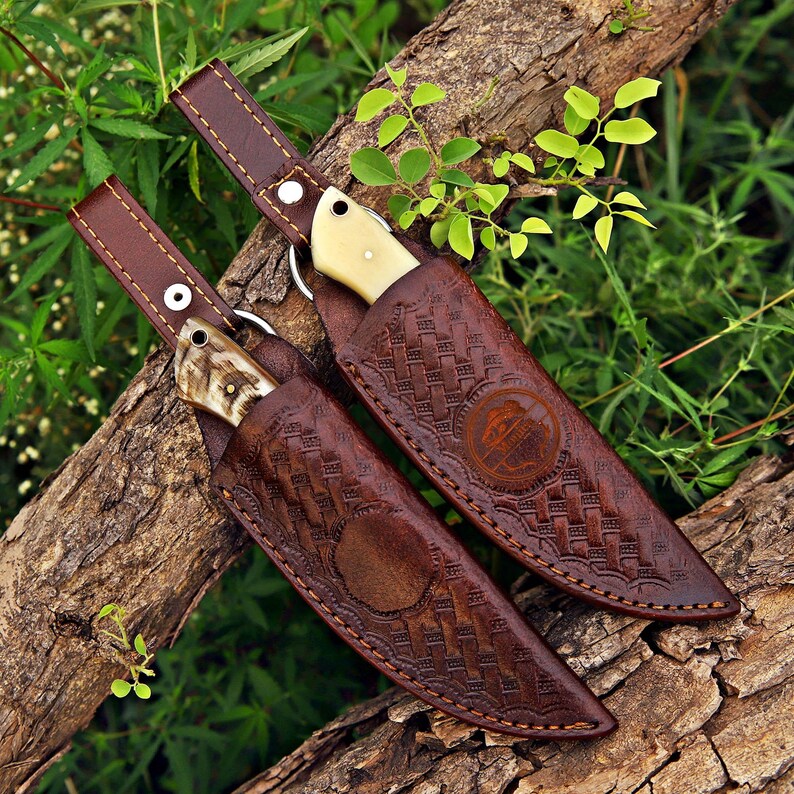 May include: Two fixed-blade knives with cream-colored handles, each housed in a dark brown leather sheath featuring an intricate basket-weave pattern, a circular embossed logo, and a long strap with a silver-toned metal snap button closure.