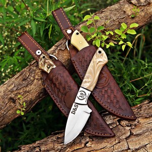 May include: Two fixed-blade knives with cream-colored handles and silver steel blades. One blade features the text D&S and a 10-year anniversary emblem. Each knife is housed in a dark brown leather sheath with embossed patterns and a belt loop.