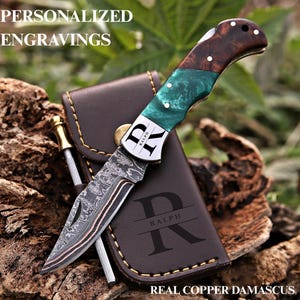 May include: A folding pocket knife with a patterned copper Damascus steel blade, a handle featuring dark wood and marbled green resin, and the name RALPH engraved on the bolster. It rests on a brown leather sheath with the letter R and RALPH.