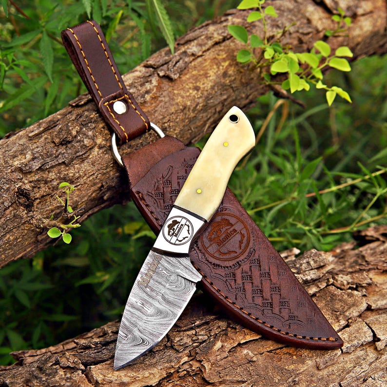 May include: A fixed-blade knife with a patterned Damascus steel blade and a smooth, cream-colored handle. The knife features a circular logo with the text "Monica" on the bolster. It is paired with a brown leather sheath featuring embossed details.