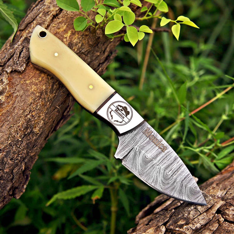 May include: A fixed-blade knife with a cream-colored handle, brass pins, and a lanyard hole. The blade features a distinct Damascus steel pattern and is engraved with the name Monica. A circular logo with a fish graphic and the name Monica is visible.