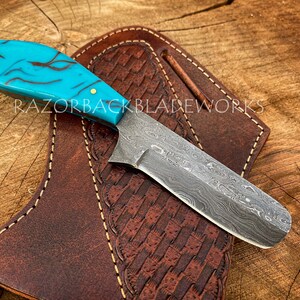 May include: A turquoise and black patterned knife with a Damascus steel blade and a leather sheath. RAZORBACKBLADEWORKS is printed on the sheath.