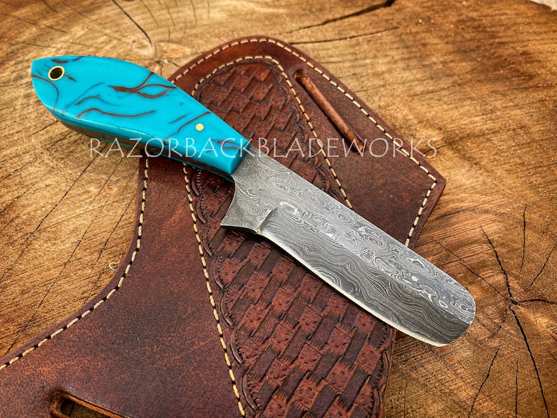 May include: A turquoise and black patterned knife with a Damascus steel blade and a leather sheath. RAZORBACKBLADEWORKS is printed on the sheath.