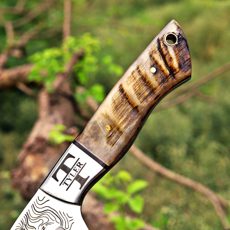 May include: A knife handle crafted from polished, variegated brown and cream horn material, featuring two small brass pins and a lanyard hole at the end. The metal bolster is engraved with the text TYLER between two stylized T-shaped symbols.