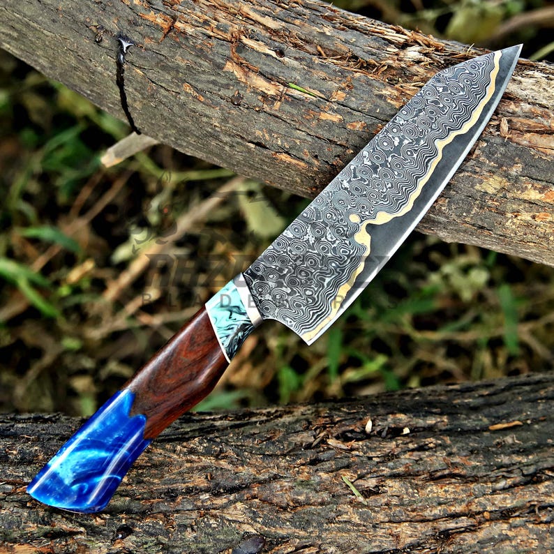 May include: A kitchen knife featuring a Damascus steel blade with a complex wavy pattern and a gold-toned edge. The handle is constructed from dark brown wood and a vibrant blue marbled resin, separated by a small section of teal and white resin.
