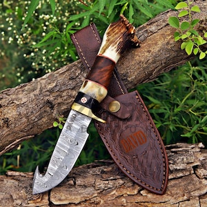 May include: A fixed-blade knife with a Damascus steel gut-hook blade engraved with the name DAVID. The handle features a dark wood section, a brass bolster with a mosaic pin, and a textured antler pommel. It includes a brown leather sheath.