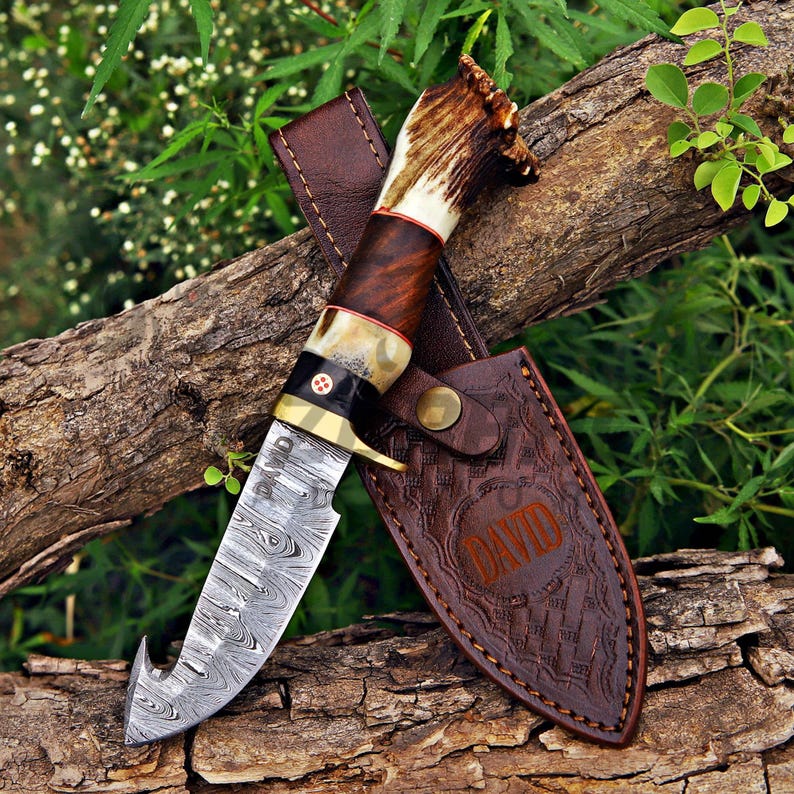 May include: A fixed-blade knife with a Damascus steel gut-hook blade engraved with the name DAVID. The handle features a dark wood section, a brass bolster with a mosaic pin, and a textured antler pommel. It includes a brown leather sheath.