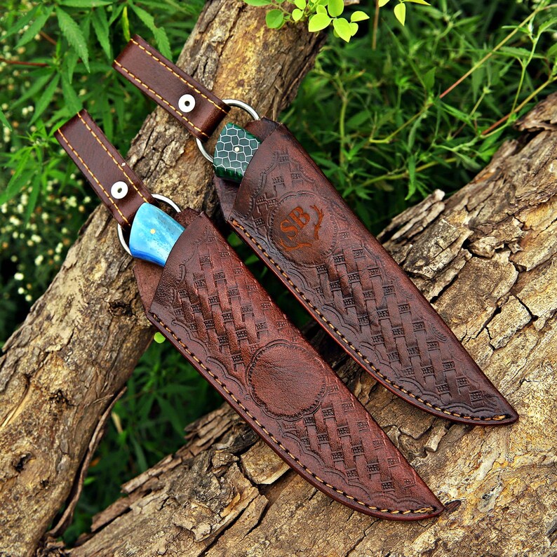 Two handcrafted fixed-blade knives partially enclosed in brown leather sheaths with basket-weave tooling, contrast stitching, and decorative embossed details, displayed on rustic tree bark outdoors.