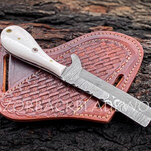 Peut inclure: Un couteau &agrave; manche blanc avec une lame en acier Damas et un fourreau en cuir. Le couteau est pos&eacute; sur une surface en bois. Le fourreau a un motif tiss&eacute; et est cousu avec du fil brun.