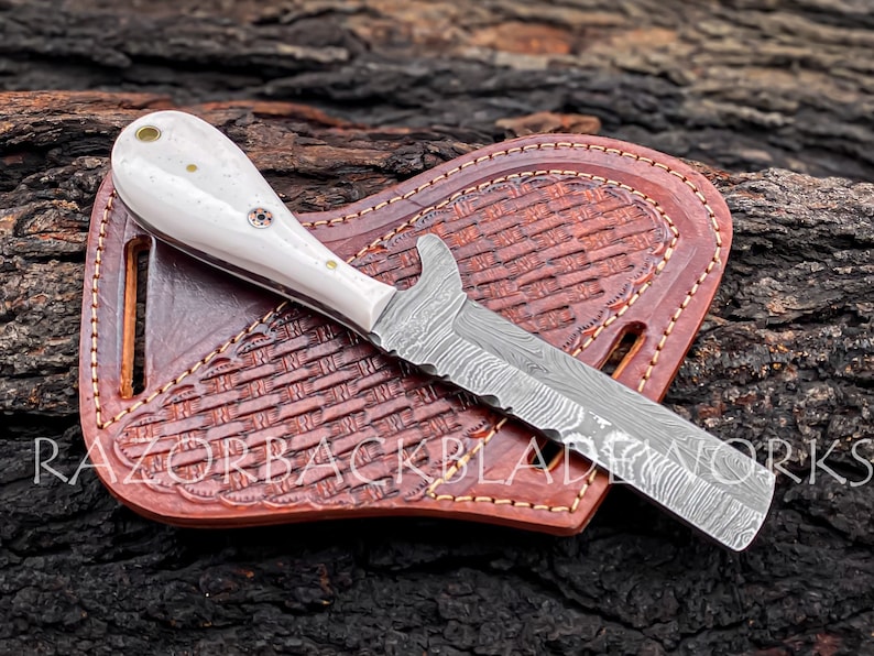 Peut inclure: Un couteau &agrave; manche blanc avec une lame en acier Damas et un fourreau en cuir. Le couteau est pos&eacute; sur une surface en bois. Le fourreau a un motif tiss&eacute; et est cousu avec du fil brun.