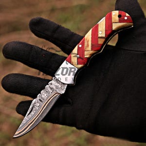 May include: A folding pocket knife featuring a Damascus steel blade with a distinct wavy pattern and a handle constructed from alternating strips of red and light tan wood arranged in a woven geometric pattern, secured with small metal rivets.