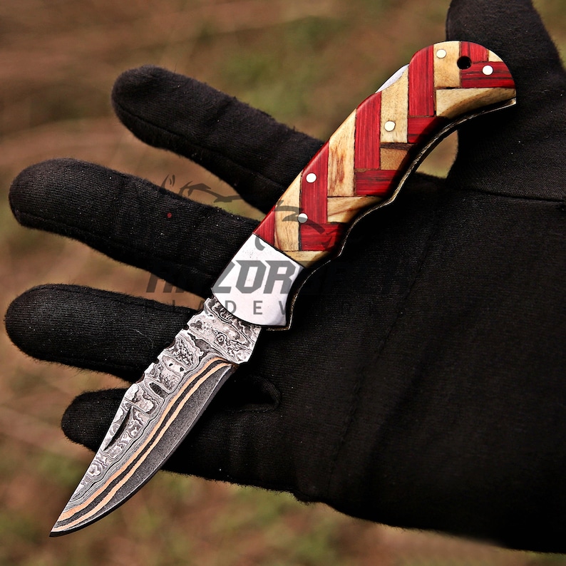 May include: A folding pocket knife featuring a Damascus steel blade with a distinct wavy pattern and a handle constructed from alternating strips of red and light tan wood arranged in a woven geometric pattern, secured with small metal rivets.