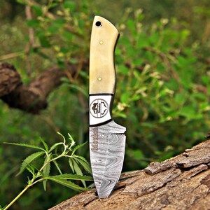 May include: A fixed-blade knife featuring a Damascus steel blade with a distinct wavy pattern and the name Monica etched on the metal. The handle is made of smooth, cream-colored material secured with two brass pins and includes a lanyard hole.