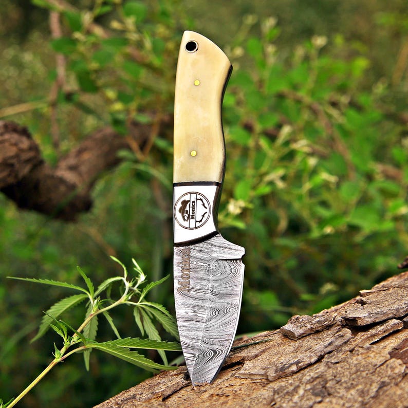 May include: A fixed-blade knife featuring a Damascus steel blade with a distinct wavy pattern and the name Monica etched on the metal. The handle is made of smooth, cream-colored material secured with two brass pins and includes a lanyard hole.