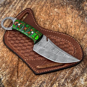 May include: A fixed-blade knife with a Damascus steel blade featuring a wavy pattern and a bright green and black layered wood handle. The handle includes a metal ring at the pommel. The knife rests on a brown leather sheath with a basket-weave pattern.