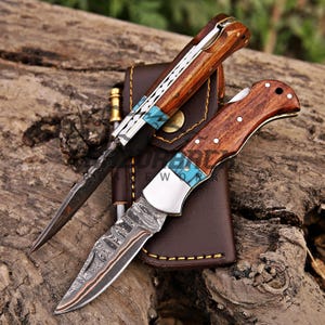 May include: Two folding pocket knives with Damascus steel blades featuring intricate patterns and wooden handles with turquoise inlays. The knives are positioned on a brown leather sheath with yellow stitching, showcasing metal bolsters and rivets.