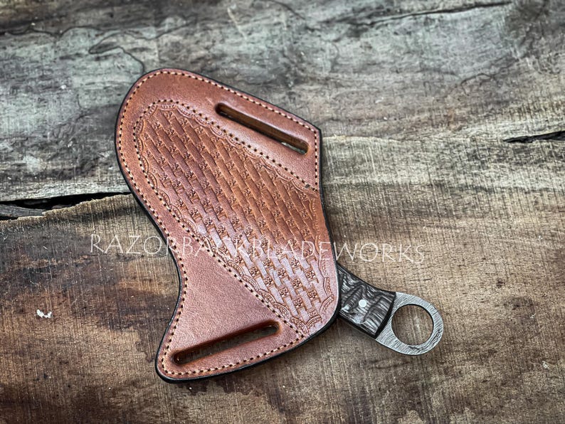 以下が含まれることがあります： バスケットウィーブ模様と2つのベルトスロットを備えたタンカラーのレザーシース。ダークな模様のハンドルとフィンガーリングポメルを持つナイフが部分的に収められている。レザーシースの中央にはRAZORBACK BLADEWORKSの文字が見える。