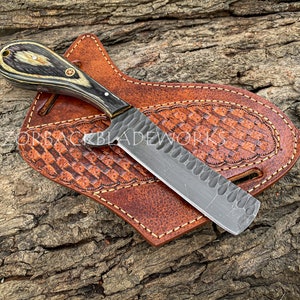 Puede incluir: Un cuchillo negro y plateado con mango de madera y funda de cuero. El cuchillo tiene una hoja texturizada y un peque&ntilde;o agujero en el mango. La funda es de cuero marr&oacute;n con un patr&oacute;n decorativo.