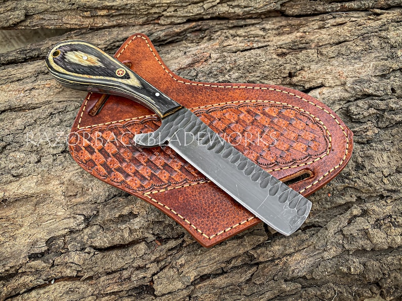 Puede incluir: Un cuchillo negro y plateado con mango de madera y funda de cuero. El cuchillo tiene una hoja texturizada y un peque&ntilde;o agujero en el mango. La funda es de cuero marr&oacute;n con un patr&oacute;n decorativo.