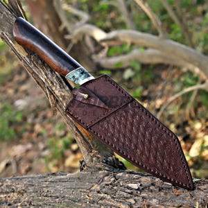 May include: A fixed-blade knife with a dark brown wooden handle and a decorative bolster featuring a marbled blue and white pattern. The blade is encased in a brown leather sheath with a textured basket-weave design and a single metal stud.