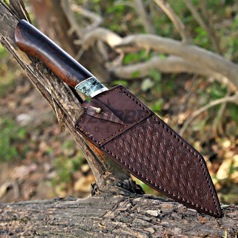 May include: A fixed-blade knife with a dark brown wooden handle and a decorative bolster featuring a marbled blue and white pattern. The blade is encased in a brown leather sheath with a textured basket-weave design and a single metal stud.