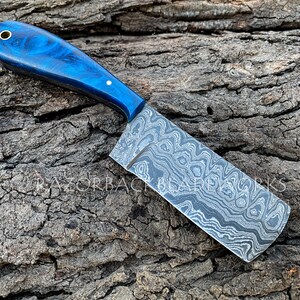 Puede incluir: Un cuchillo de chef con patr&oacute;n azul y negro con mango de madera azul. El cuchillo est&aacute; sobre una superficie de madera.