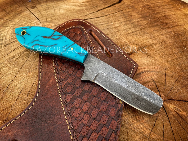 May include: A turquoise and black patterned handle knife with a Damascus steel blade. The knife is resting on a brown leather sheath with decorative stitching.