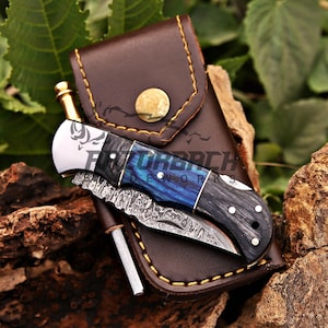 May include: A folding pocket knife with a Damascus steel blade, a blue and black wood handle, and a silver bolster featuring a boar engraving. The knife rests on a brown leather sheath with yellow stitching and a brass snap button closure.