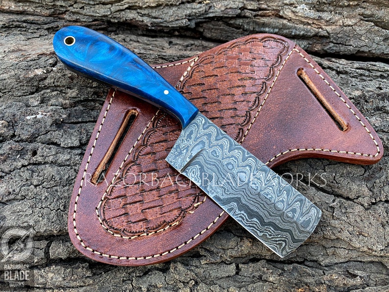 Peut inclure: Un couteau &agrave; motif bleu et argent avec un manche bleu et un fourreau en cuir. Le couteau est sur une surface en bois.