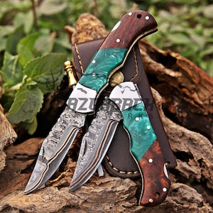 May include: Three folding knives with patterned blades and wooden handles with green and brown accents. The knives are displayed with a brown leather sheath on a wooden surface.