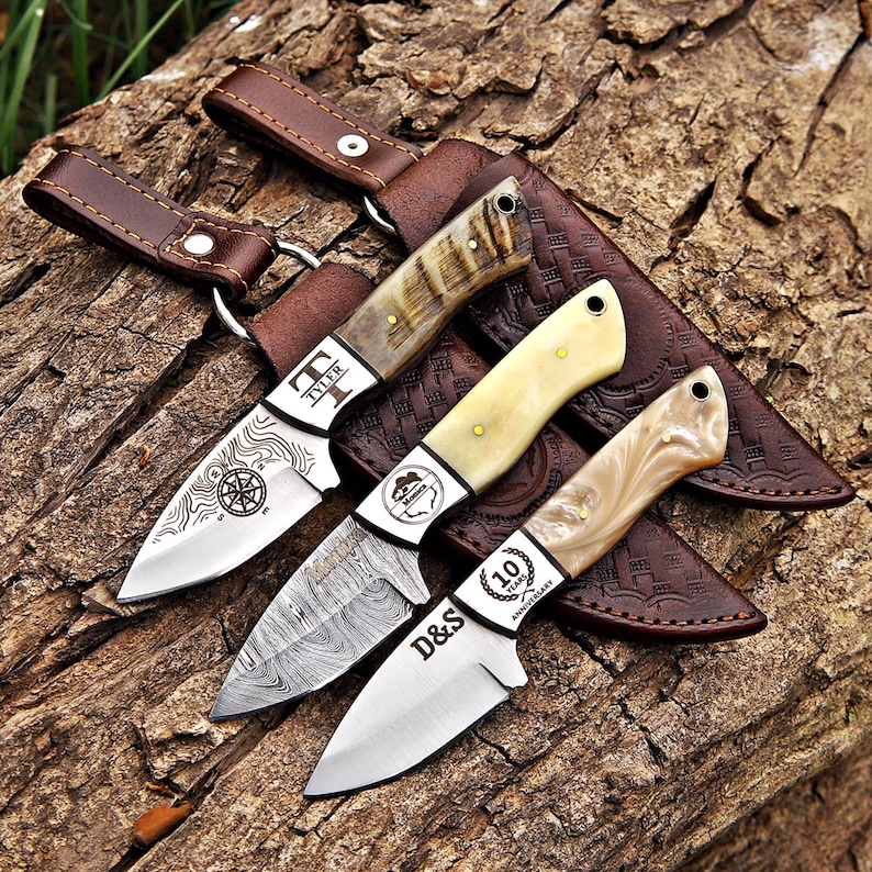 May include: Three fixed-blade knives with distinct handles and leather sheaths. The top knife has a horn handle and a compass engraving. The middle knife features a Damascus steel blade and a cream handle. The bottom knife has a marbled handle and text reading D&S 10 Years Anniversary.