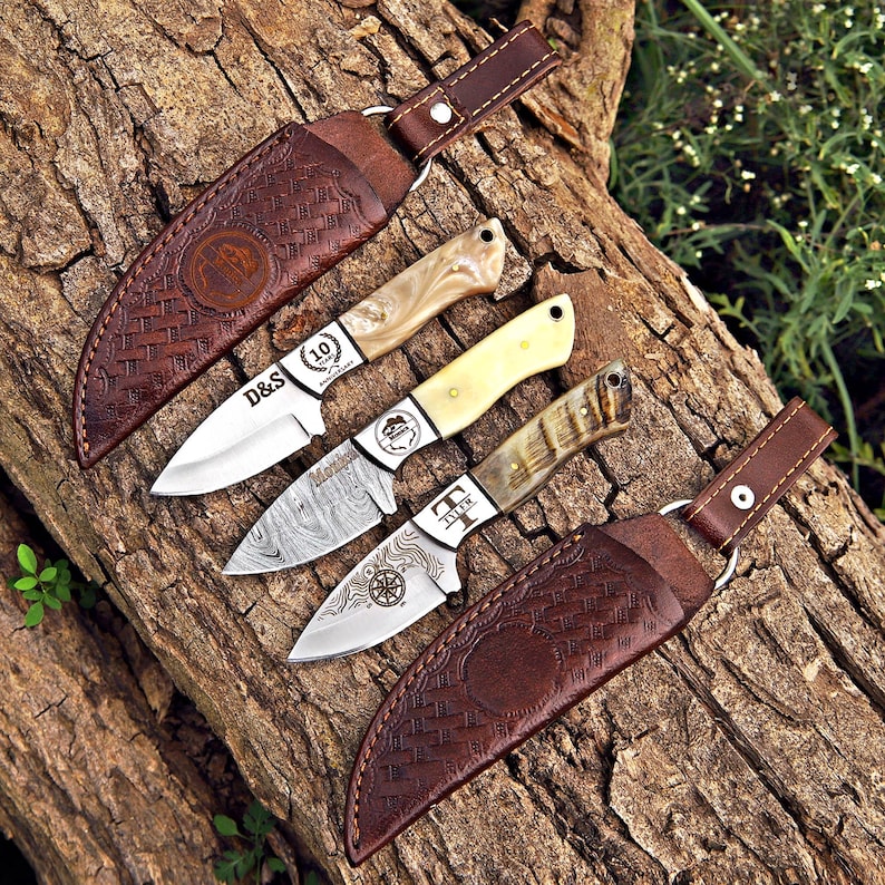 May include: Three fixed-blade knives with distinct handles and two brown leather sheaths with woven patterns. The top knife has a pearlescent handle and D&S 10th Anniversary text. The middle knife has a cream handle, and the bottom knife has a textured handle with a compass graphic and the name Tyler.