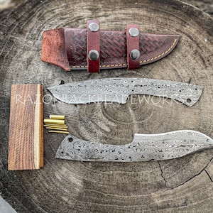 Peut inclure: Deux lames de couteau en acier Damas non finies avec un manche en bois et des broches en laiton. Les lames sont sur une surface en bois avec un fourreau en cuir.