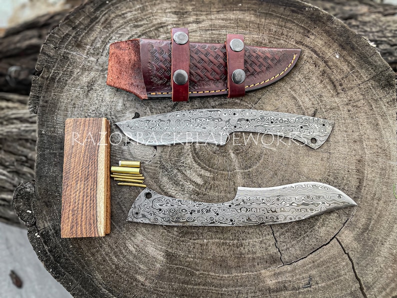 Peut inclure: Deux lames de couteau en acier Damas non finies avec un manche en bois et des broches en laiton. Les lames sont sur une surface en bois avec un fourreau en cuir.