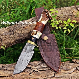 May include: A fixed-blade knife with a Damascus steel blade, a brass bolster, and a handle crafted from dark wood and textured antler. The knife is accompanied by a brown leather sheath featuring a stamped basket-weave pattern and a metal snap.