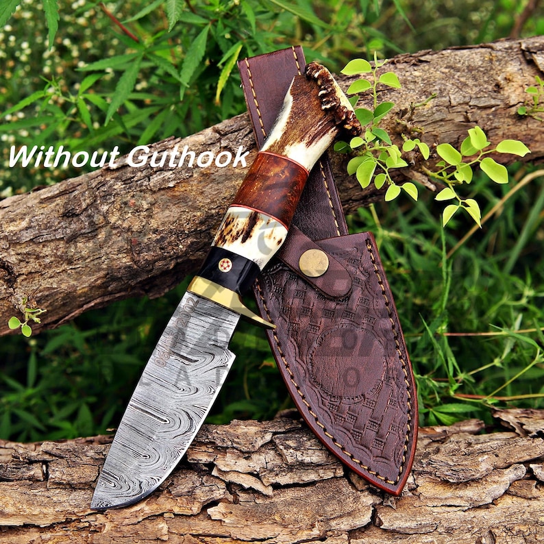 May include: A fixed-blade knife with a Damascus steel blade, a brass bolster, and a handle crafted from dark wood and textured antler. The knife is accompanied by a brown leather sheath featuring a stamped basket-weave pattern and a metal snap.