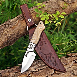 May include: A fixed-blade knife with a polished steel blade featuring the text D&S and a 10 Years Anniversary emblem. The handle is made of a cream-colored marbled material. It includes a brown leather sheath with embossed geometric patterns.
