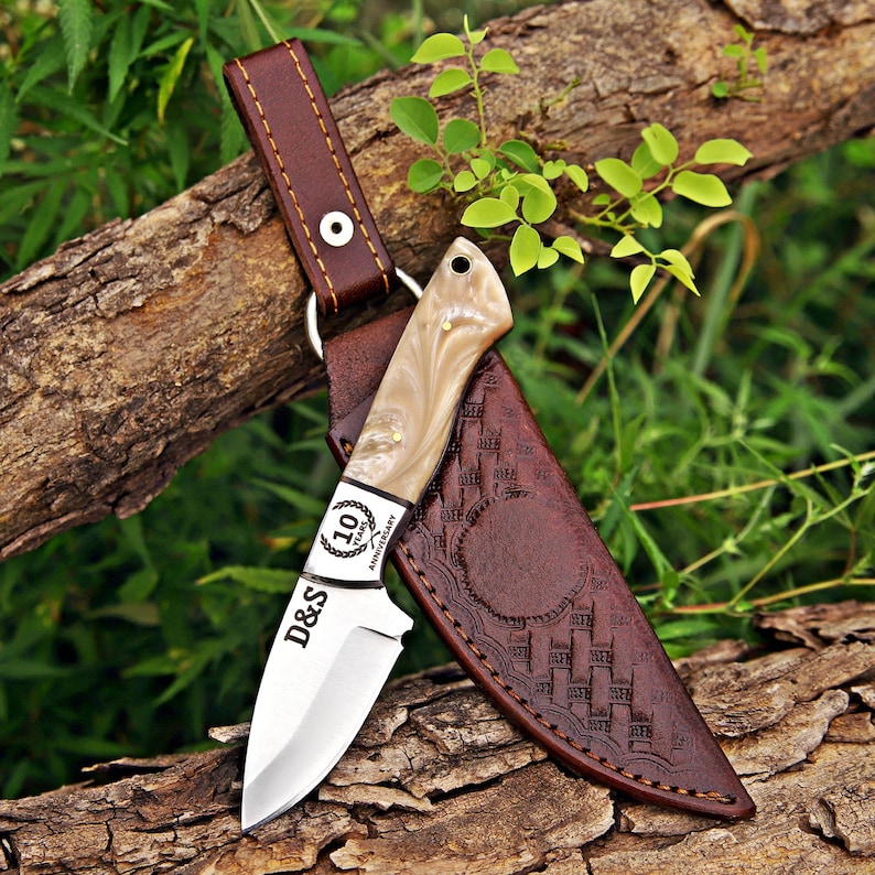 May include: A fixed-blade knife with a polished steel blade featuring the text D&S and a 10 Years Anniversary emblem. The handle is made of a cream-colored marbled material. It includes a brown leather sheath with embossed geometric patterns.