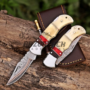 Op de afbeelding: Twee inklapbare zakmessen met Damascus stalen bladen voorzien van een koperen inleg. De handvatten zijn gemaakt van cr&egrave;mekleurig bot, een rood accentgedeelte en donker hout, vastgezet met metalen pinnen. Een bruine lederen schede is inbegrepen.