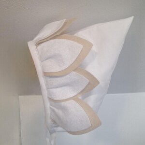 May include: A white bonnet featuring three layered petal-shaped details in off-white and beige. The bonnet has a pointed top and ties with a white ribbon. The design is simple and elegant, suitable for a baby.