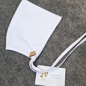 May include: A white baby bonnet with a wooden button and long white ties. The bonnet has a rounded shape and is made of a soft fabric. A tag with the brand name "Pearl & May" is attached.