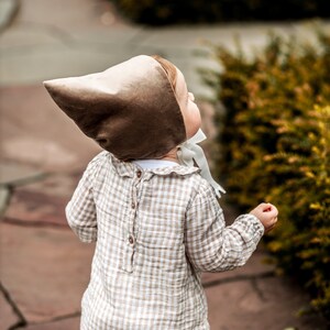 May include: A toddler wearing a brown velvet bonnet with a white ribbon and a beige and white gingham romper.