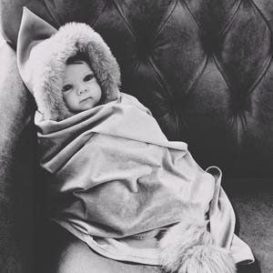 May include: A baby wrapped in a soft, light grey blanket and hooded garment with a faux fur trim. The baby is seated on a tufted, dark grey upholstered surface.