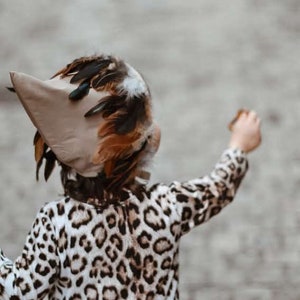 May include: A child wearing a brown and white leopard print dress with a brown and white feathered hat. The hat has a long beak and is shaped like a bird's head.