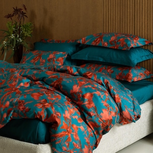 May include: A teal and orange floral patterned bedding set on a bed with a light grey upholstered frame. The set includes a duvet cover, fitted sheet, and several pillows. The background is a brown wooden wall.