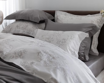 May include: A bed with gray and white bedding. The pillows and duvet feature a floral pattern in shades of gray. The bed is made with a gray fitted sheet and several gray pillows. The overall aesthetic is soft and inviting.