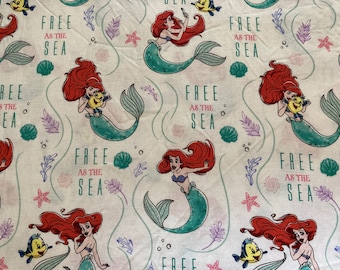 May include: A white fabric with a repeating pattern of Ariel, the Little Mermaid, holding a seashell and a fish. The text "Free as the Sea" is repeated throughout the fabric.