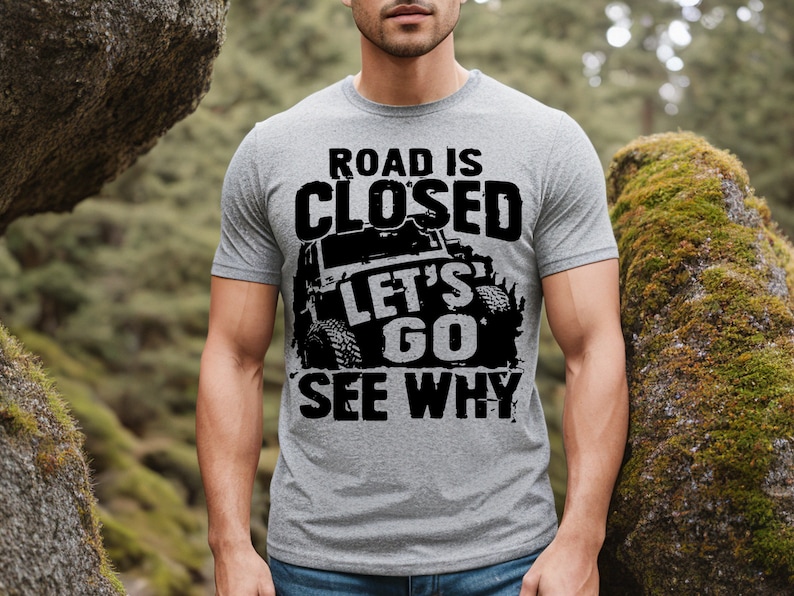 May include: A gray t-shirt with the text "ROAD IS CLOSED LET'S GO SEE WHY" in a distressed black font. The graphic includes a black off-road vehicle. The shirt is short-sleeved and worn by a person.